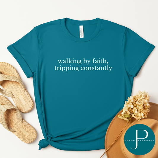 Flat lay of a marine blue t-shirt with the words “walking by faith, tripping constantly” in simple white text. Styled with neutral flip-flop sandals, a tan felt hat, and dried flowers.